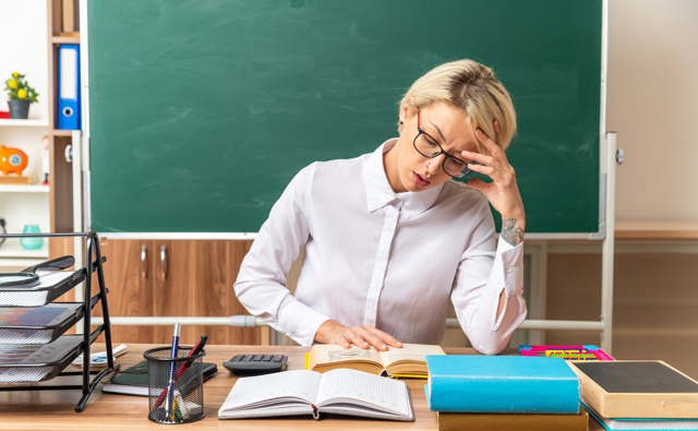 Concentrated Young Blonde Female Teacher Wearing Glasses Sitting Desk With School Tools Classroom Keeping Hand Head Open Book Reading Book